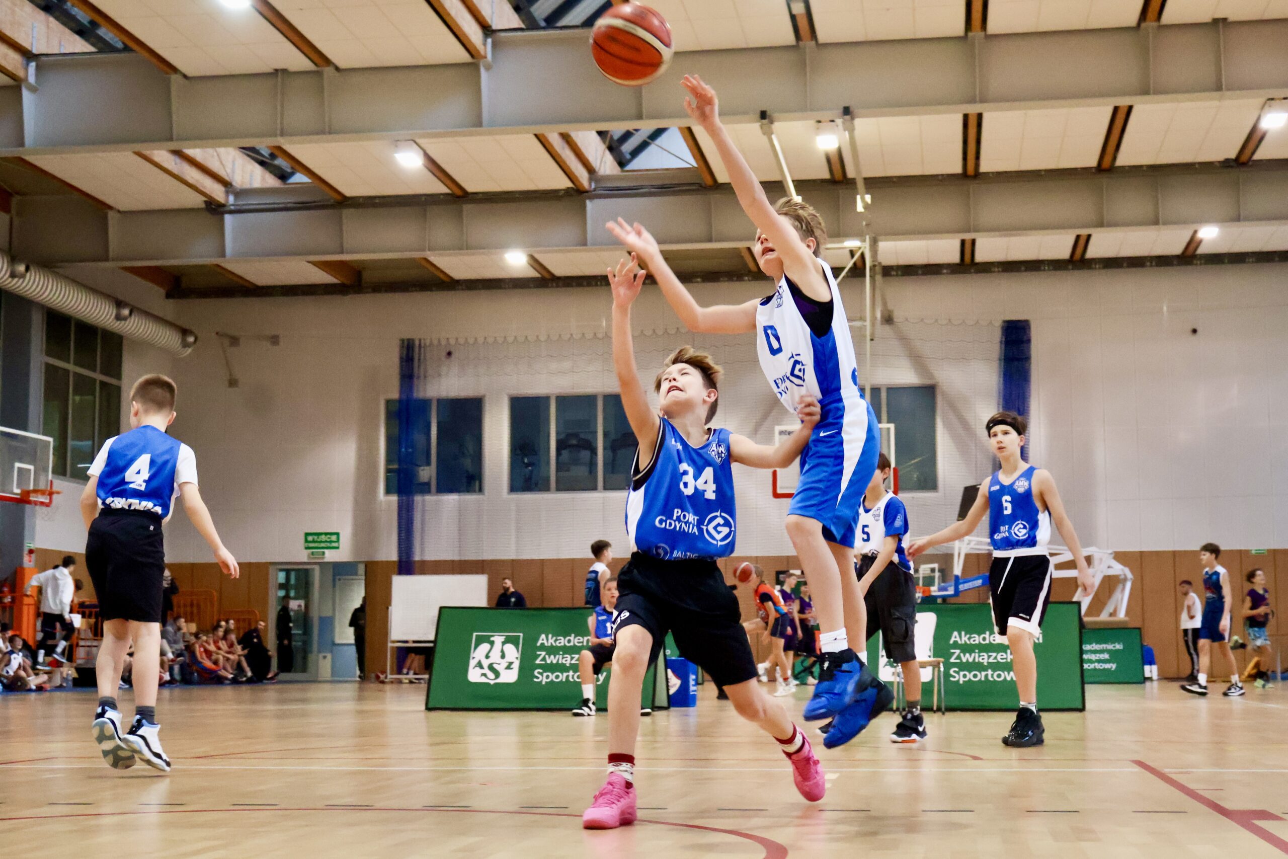 Chłopiec w białej koszulce oddaje rzut do kosza, a obrońca w niebieskiej koszulce próbuje go zablokować.