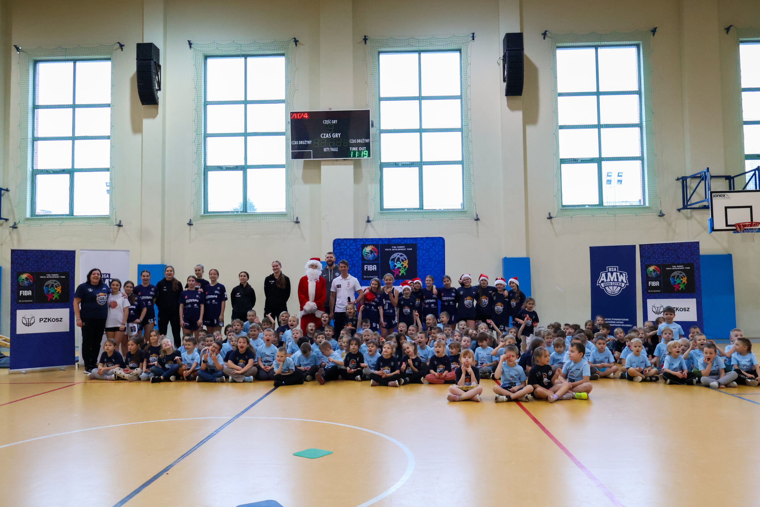 Zdjęcie grupowe uczestników wydarzenia sportowego – dzieci i trenerzy w niebieskich strojach pozują w hali sportowej.