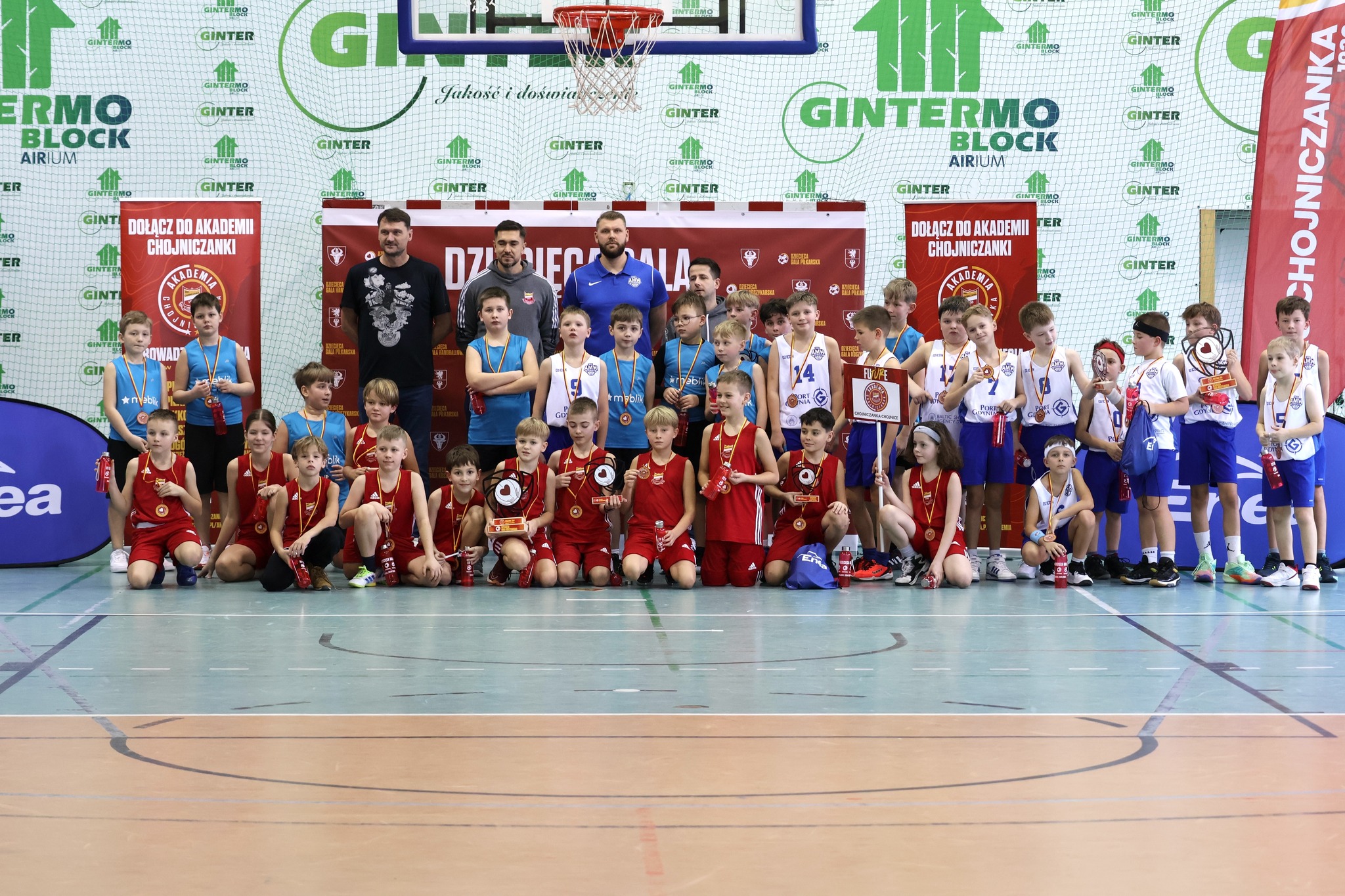 Duża grupa młodych koszykarzy z różnych drużyn pozuje do wspólnego zdjęcia na tle tablicy w hali sportowej, po turnieju u11.
