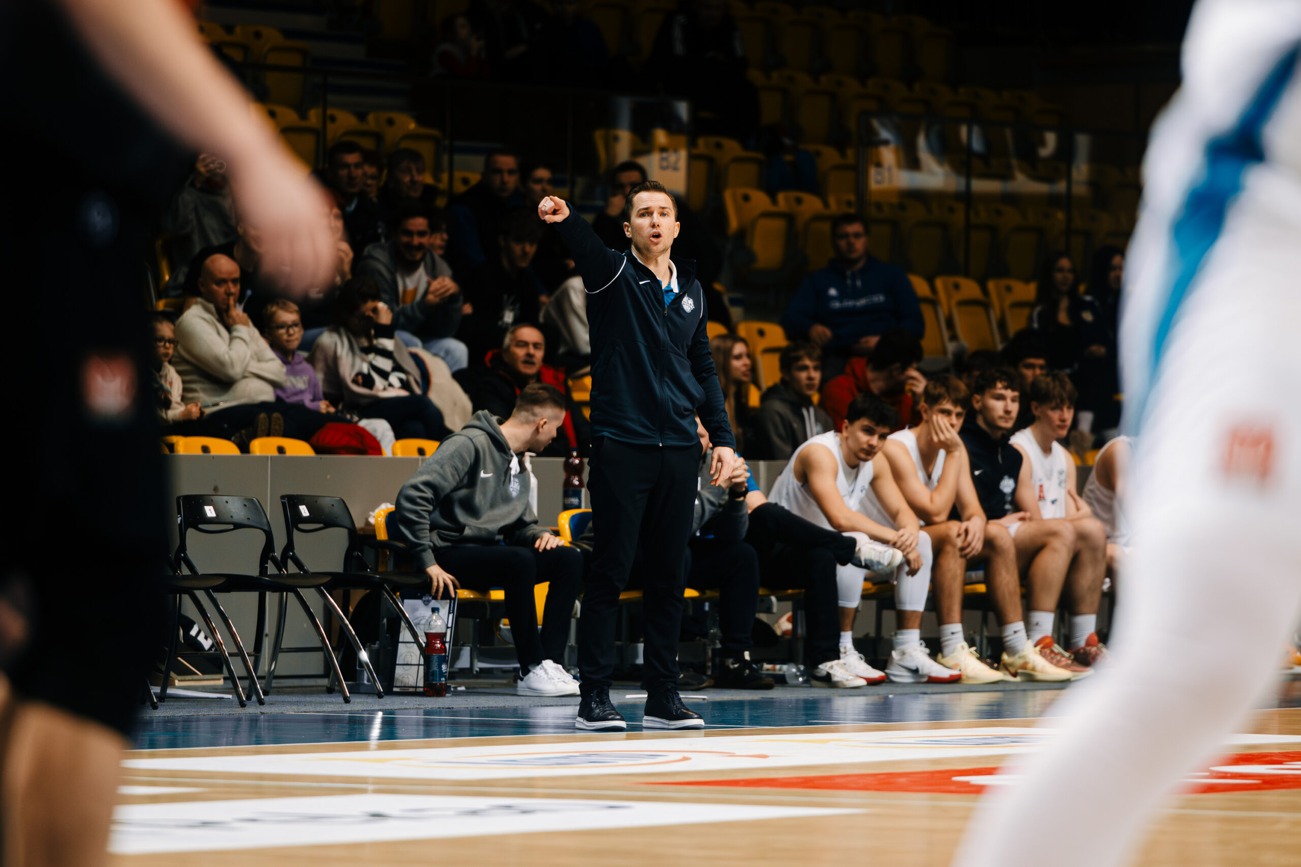 Trener w granatowej bluzie intensywnie gestykuluje przy linii bocznej, prowadząc swój zespół.