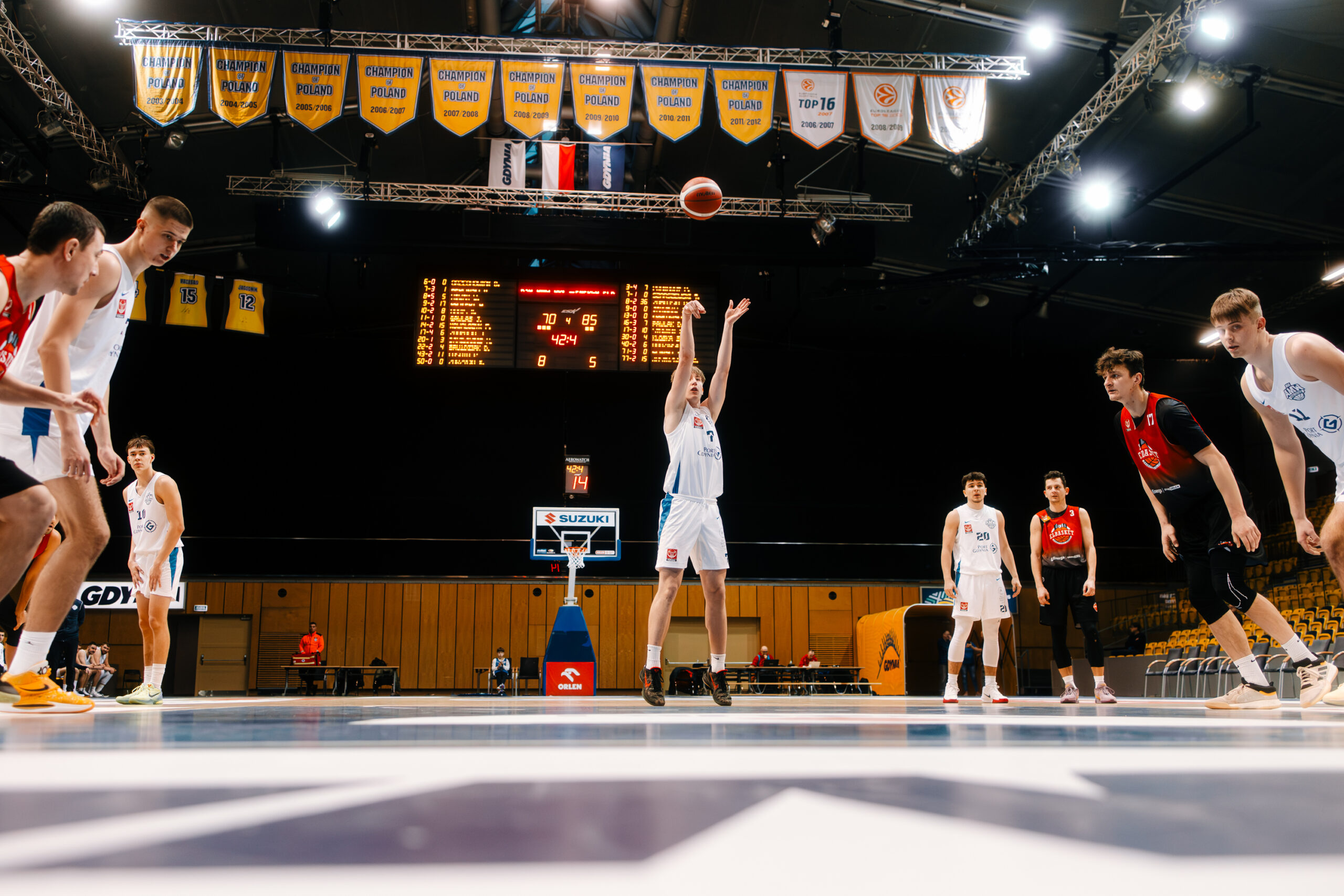 Koszykarz na linii rzutów wolnych, w pełnym skupieniu oddaje rzut, podczas gdy w tle widać tablicę wyników i zawieszone nad boiskiem banery.