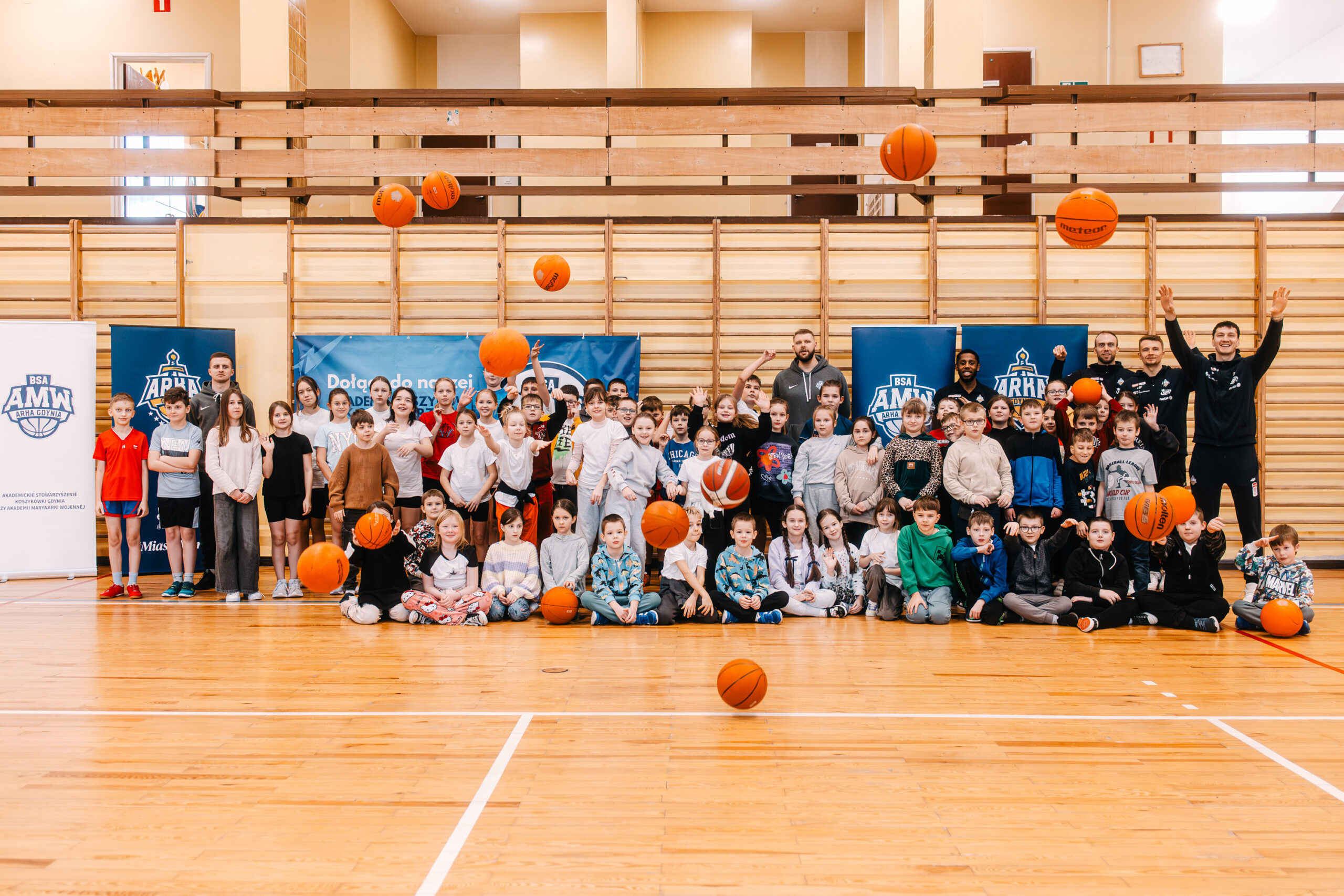 Dzieci rzucają piłki do koszykówki w górę podczas wspólnego zdjęcia w hali sportowej.