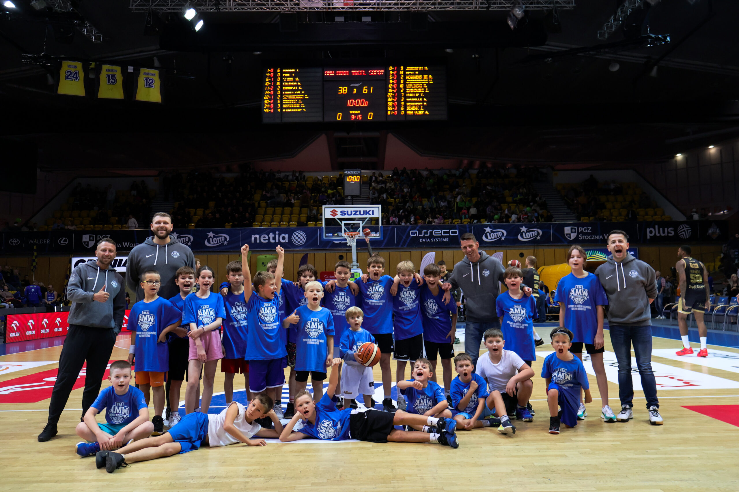 Grupa młodych koszykarzy w niebieskich koszulkach pozuje do zdjęcia na środku boiska, za nimi trybuny oraz tablica wyników.