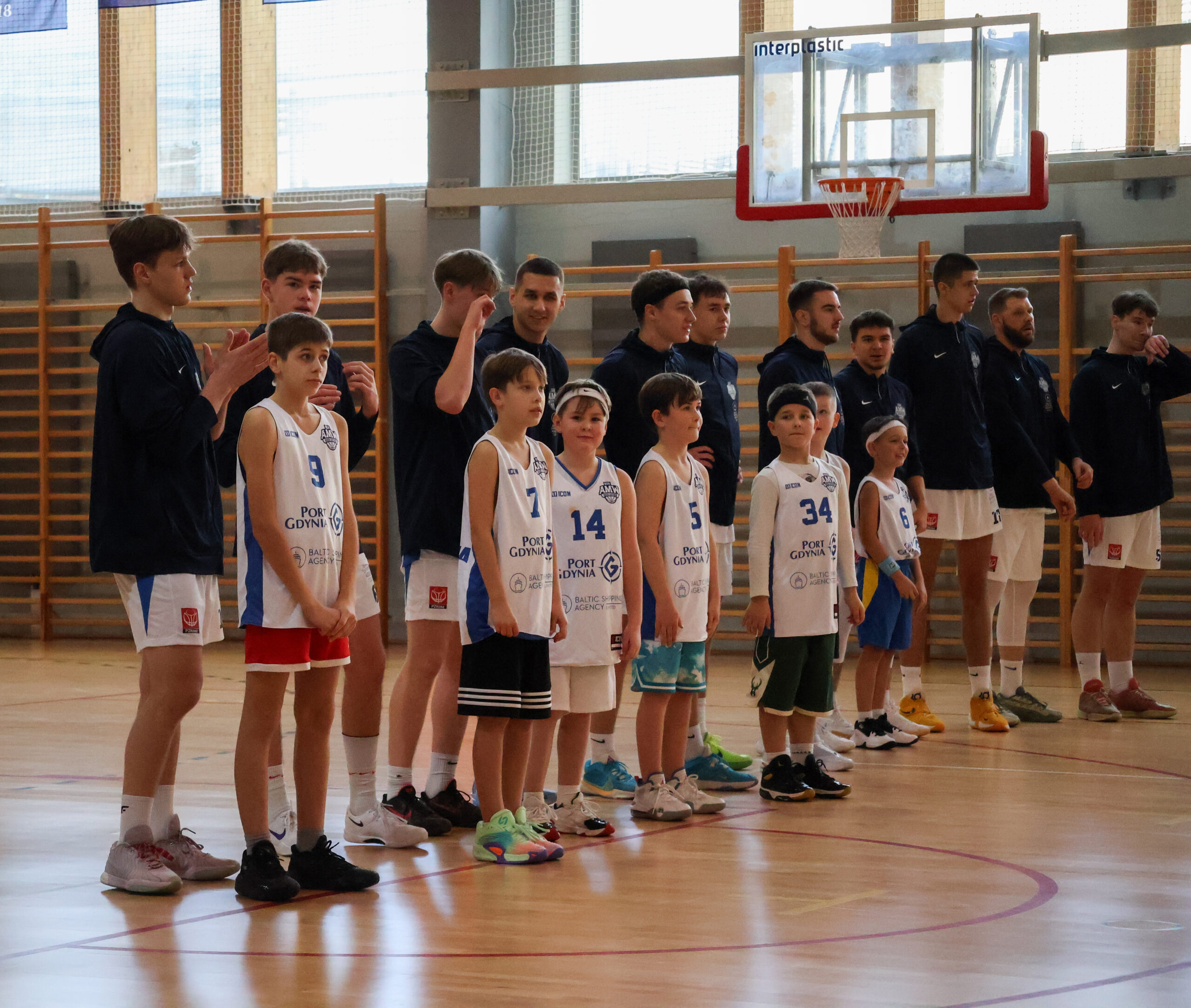 Młodzi zawodnicy ustawieni w rzędzie na boisku, czekający na rozpoczęcie gry wraz ze starszymi zawodnikami 2 ligi.