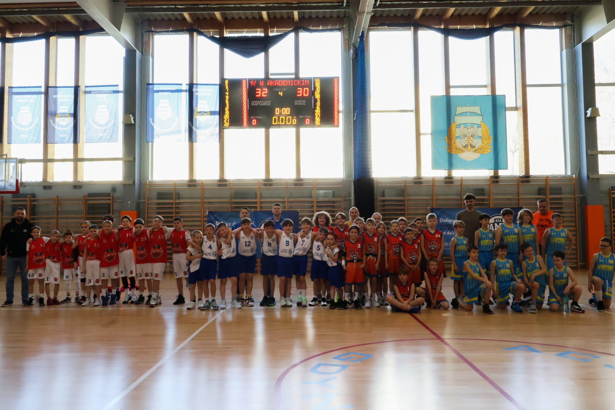 Wspólne zdjęcie kilku drużyn w różnych kolorowych strojach na tle tablicy wyników.