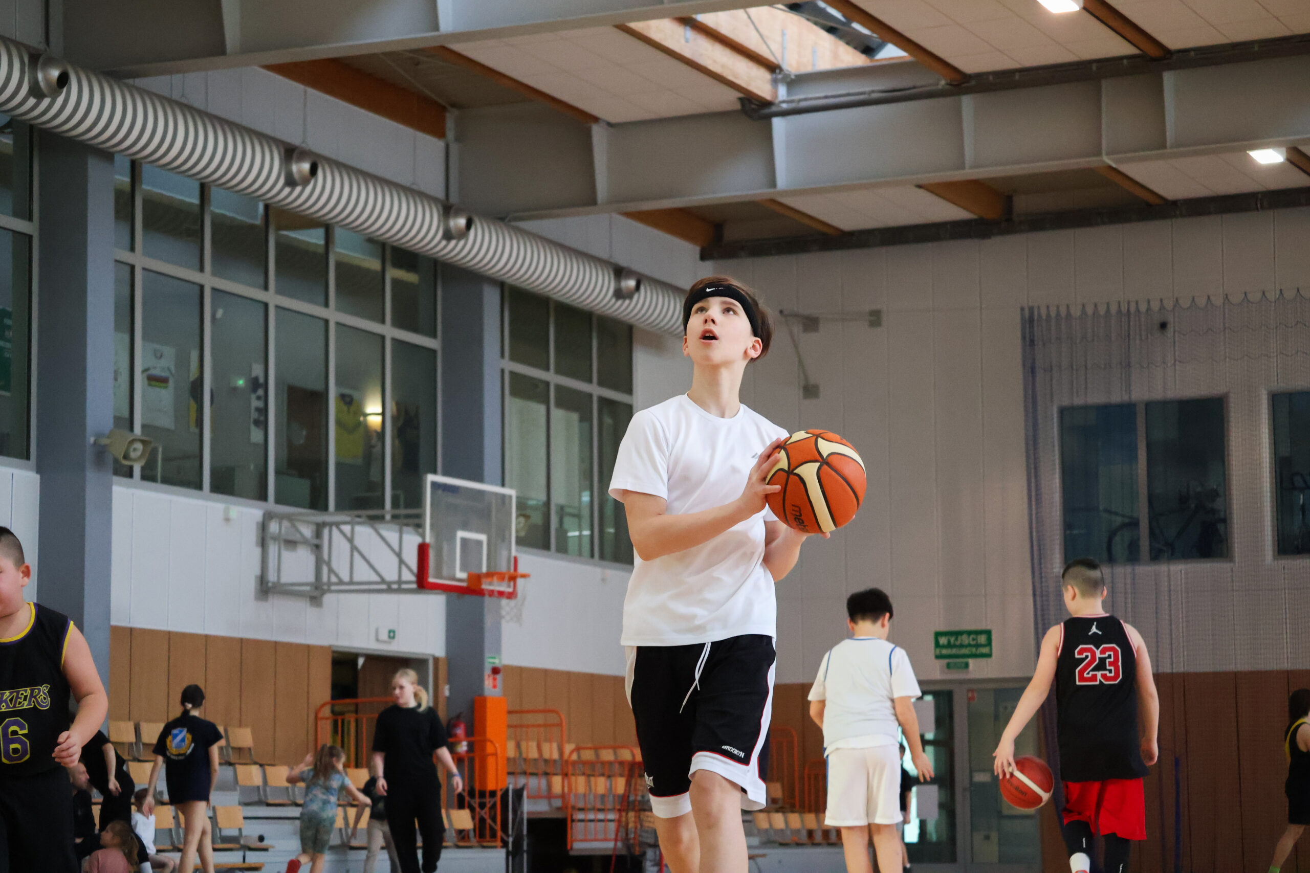 – Chłopiec w białej koszulce i czarnych spodenkach wykonuje rzut do kosza na hali sportowej. W tle inni uczestnicy treningu.