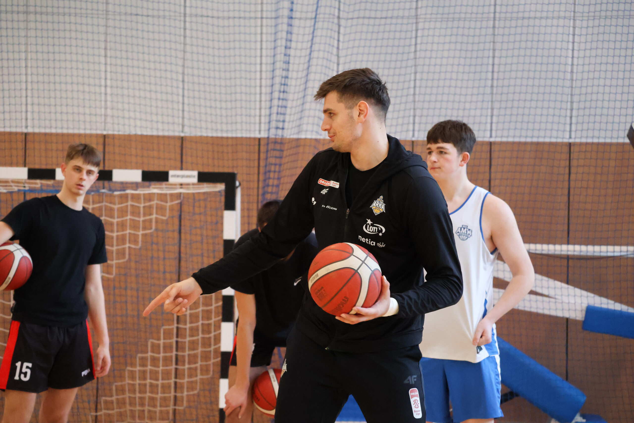 Stefan Djordevic - zawodnik ekstraklasy w czarnym dresie instruuje innych zawodników, trzymając piłkę do koszykówki w jednej ręce.