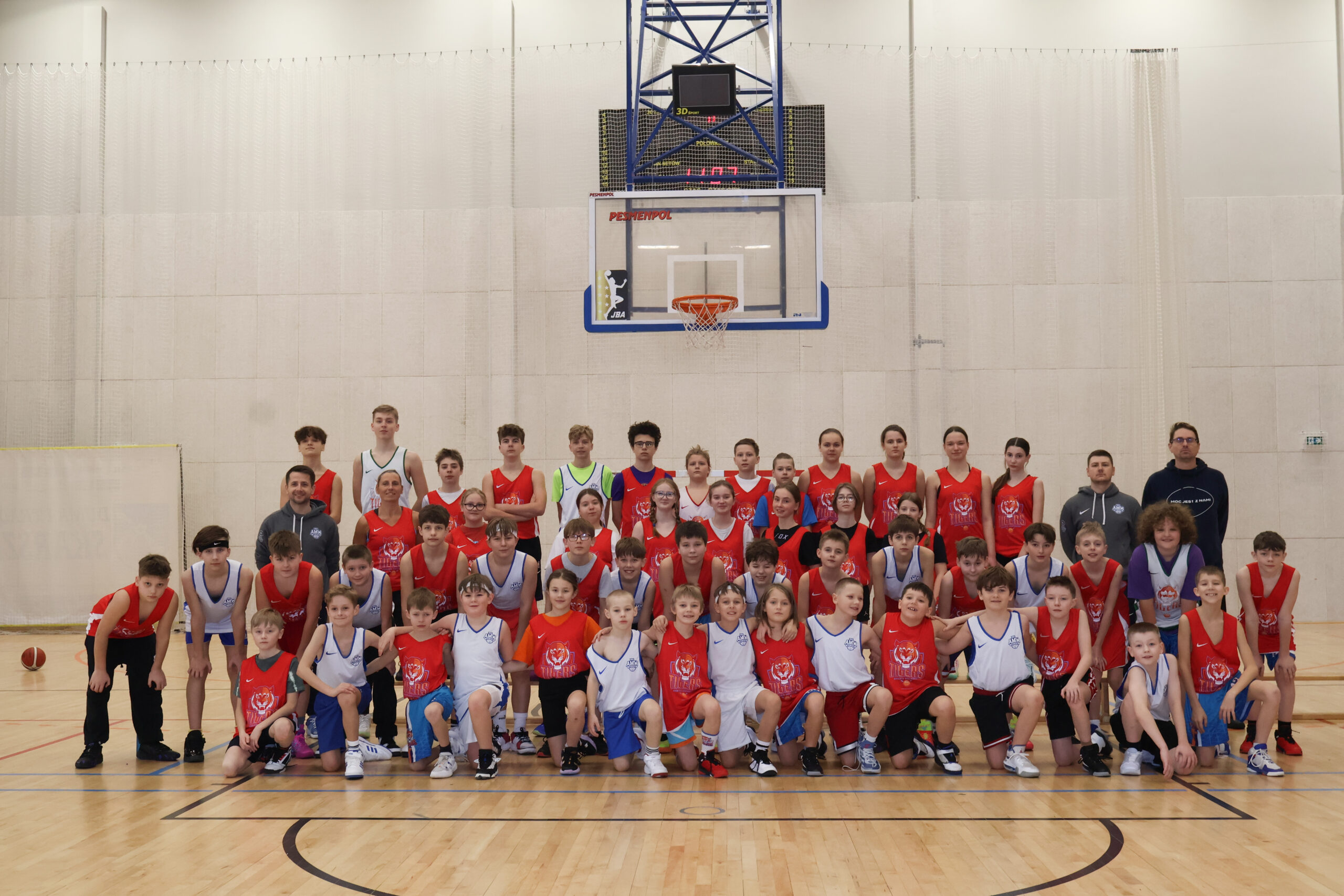 Duża grupa młodych koszykarzy w białych i czerwonych strojach pozuje do wspólnego zdjęcia pod koszem na hali sportowej.