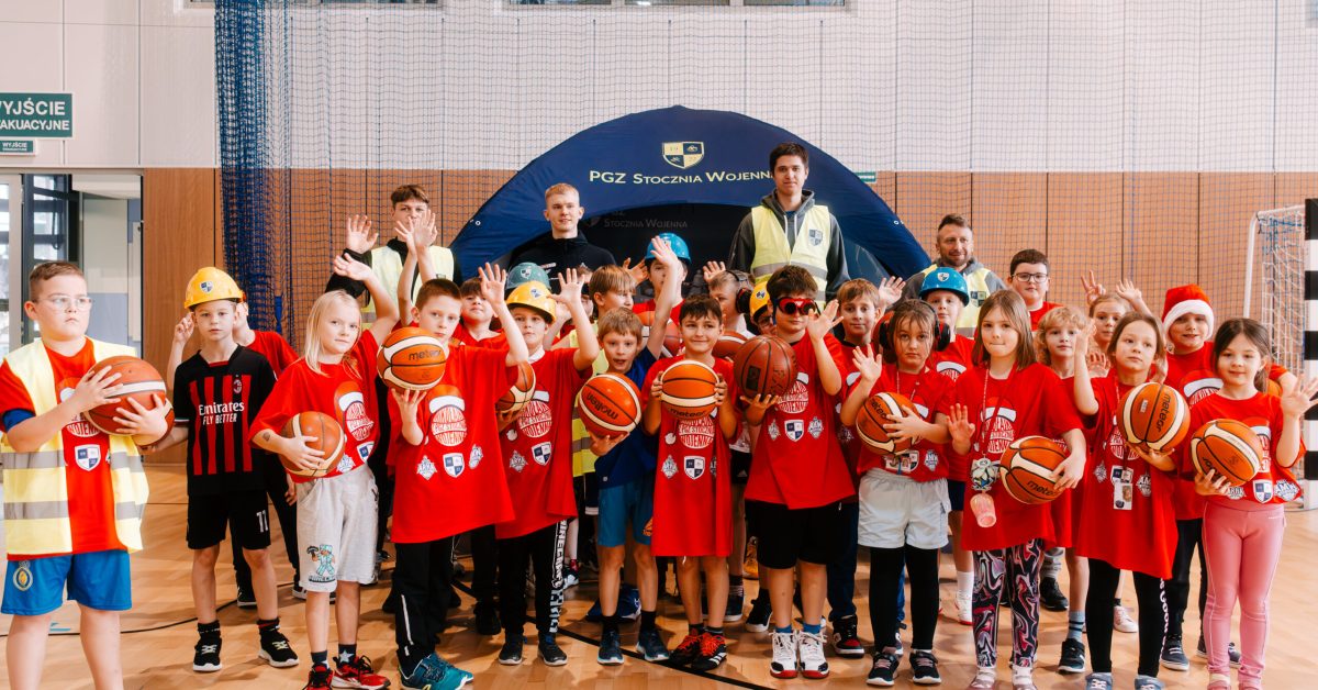 Grupa dzieci w kaskach budowlanych i kamizelkach odblaskowych pozuje do zdjęcia w hali sportowej.