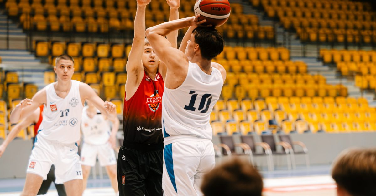 Zawodnik z numerem 10 oddaje rzut, podczas gdy obrońca stara się go utrudnić, podnosząc ręce do bloku.