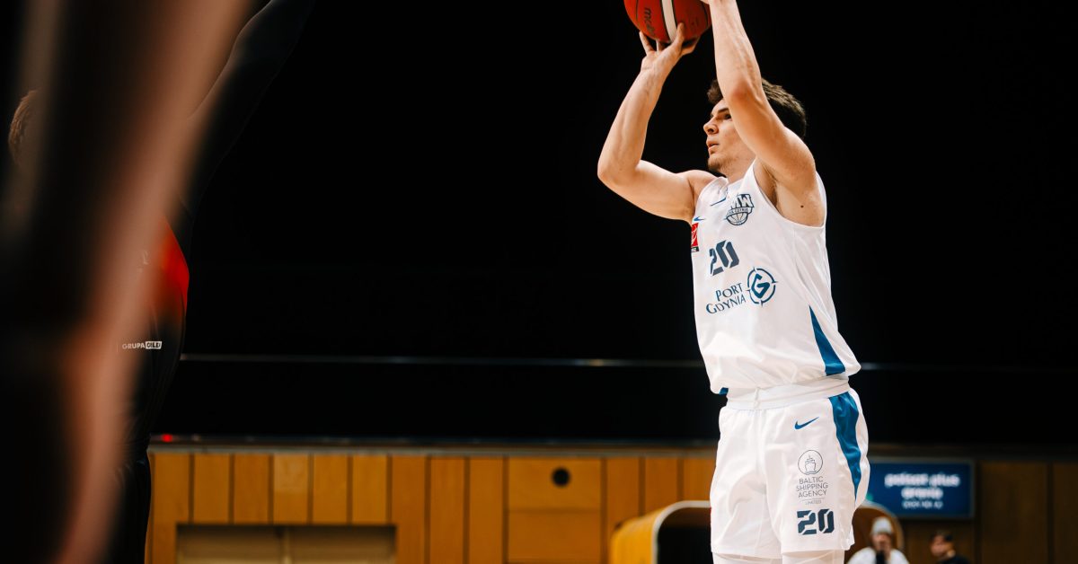 Koszykarz w białej koszulce wykonuje rzut z wyskoku, trzymając piłkę oburącz nad głową, a jego sylwetka jest wyraźnie zarysowana na tle ciemnej hali sportowej.