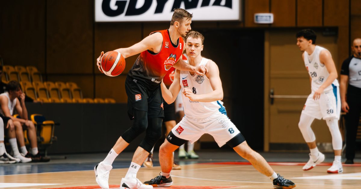 Dwóch koszykarzy w starciu na parkiecie; zawodnik w czerwonej koszulce broni piłki, podczas gdy gracz w białej koszulce próbuje bronić.