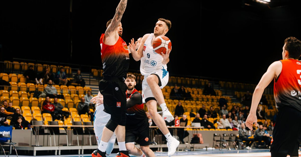 Zawodnik w białej koszulce wykonuje rzut, podczas gdy obrońca w czerwono-czarnym stroju próbuje go zablokować, wyskakując w górę.