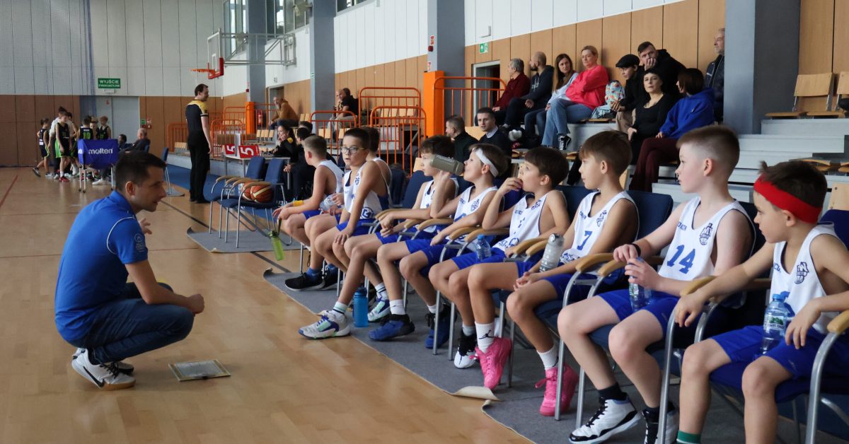 Ławka z młodymi koszykarzami w białych strojach obserwuje trenera, który daje wskazówki na meczu na hali sportowej.