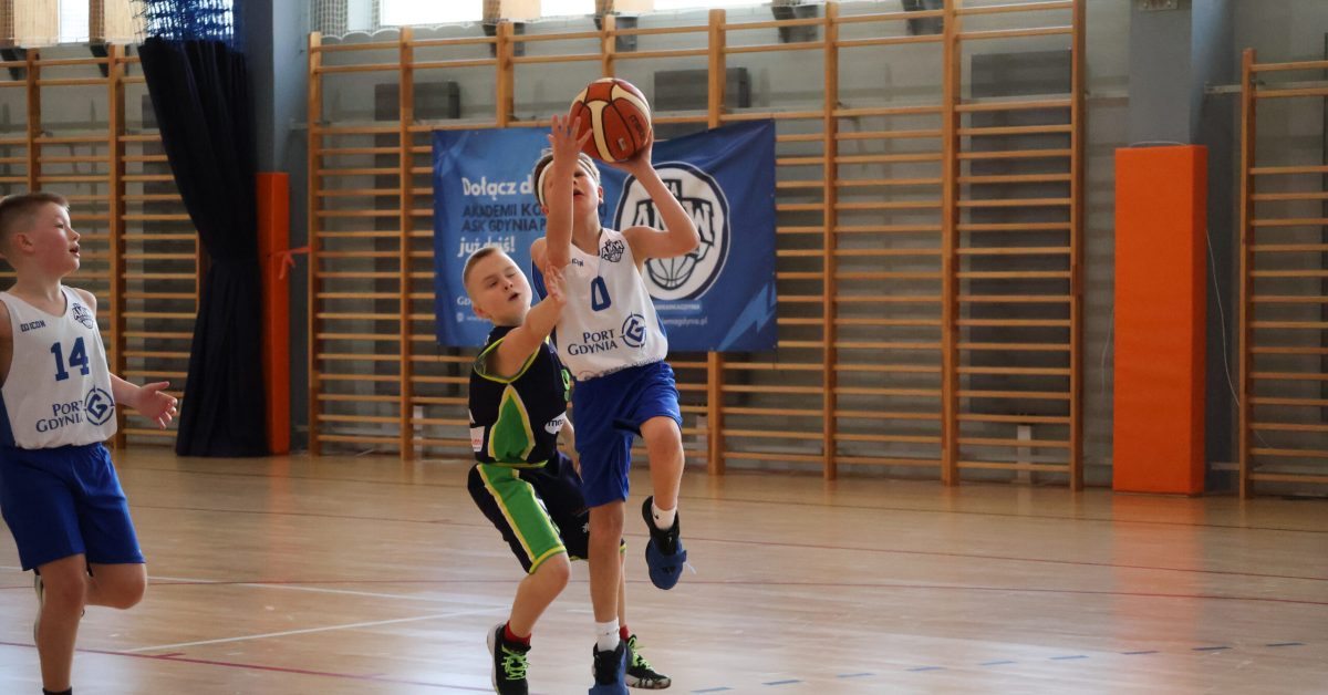 Młody koszykarz w białej koszulce oddaje rzut do kosza, podczas gdy obrońca w zielonym stroju biegnie i blokuje go obok.