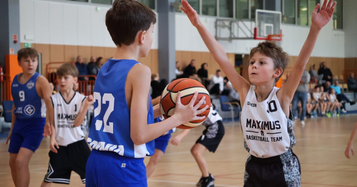 Młody koszykarz w niebieskiej koszulce z numerem 22 przygotowuje się do podania, podczas gdy obrońca w czarno-białym stroju unosi rękę do bloku.