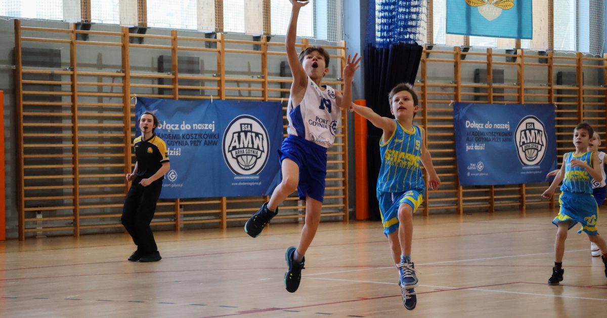 Młody koszykarz w białym stroju wyskakuje wysoko, próbując oddać rzut do kosza.