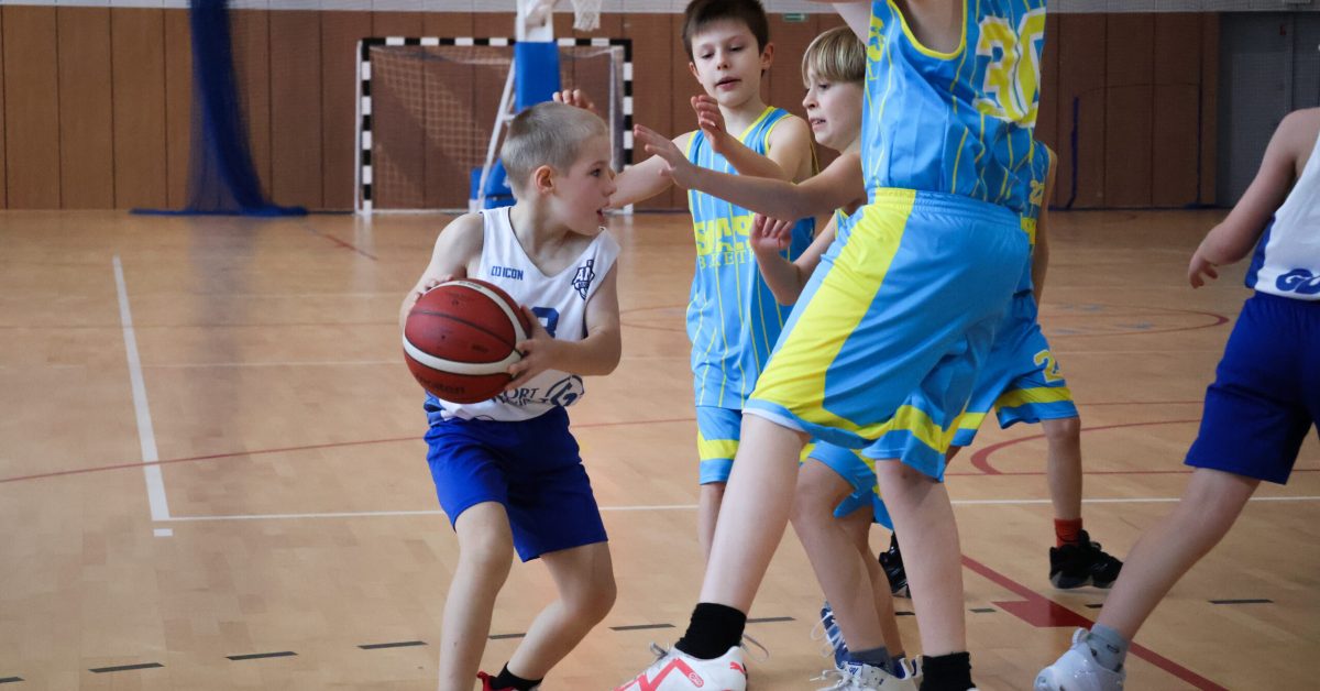 Młody zawodnik w białej koszulce i niebieskich spodenkach próbuje przedrzeć się przez obronę przeciwników w żółto-niebieskich strojach, podczas turnieju u11.