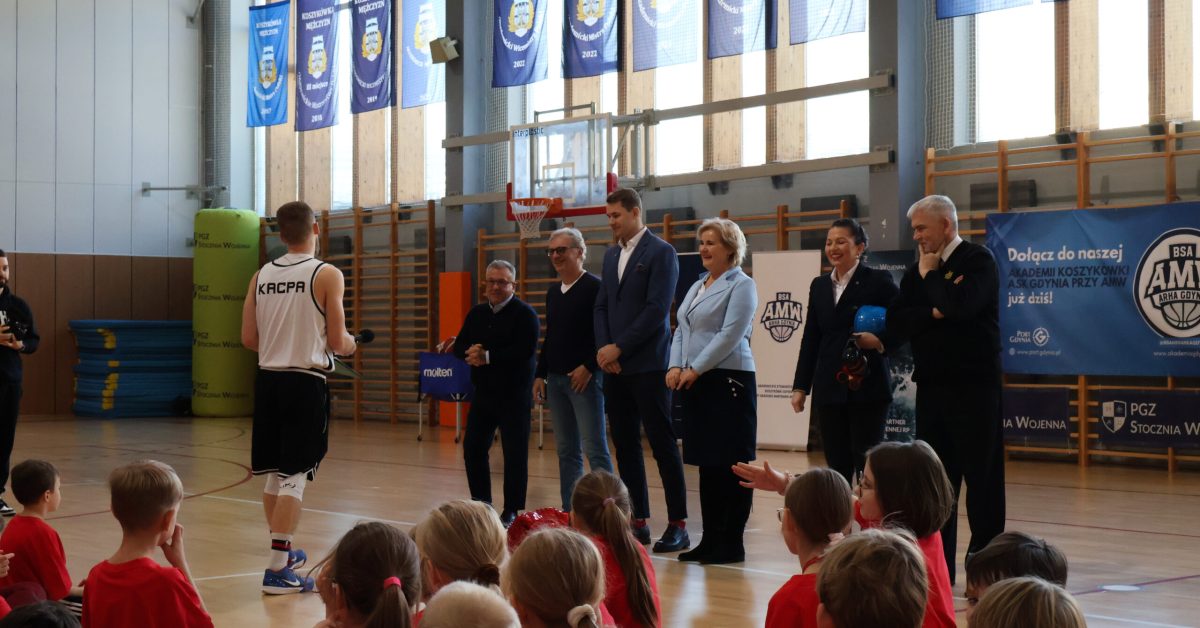 Grupa dorosłych, w tym trener i osoby w garniturach, stoi na boisku, przemawiając do dzieci siedzących na podłodze.