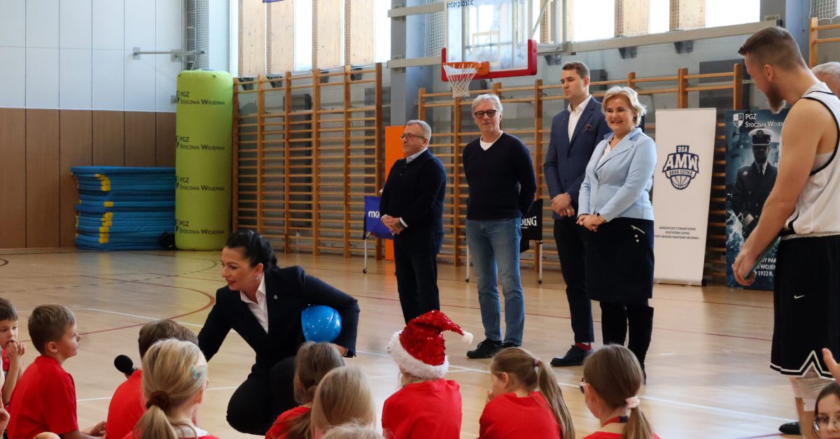 Grupa dorosłych, w tym trener i osoby w garniturach, stoi na boisku, przemawiając do dzieci siedzących na podłodze.