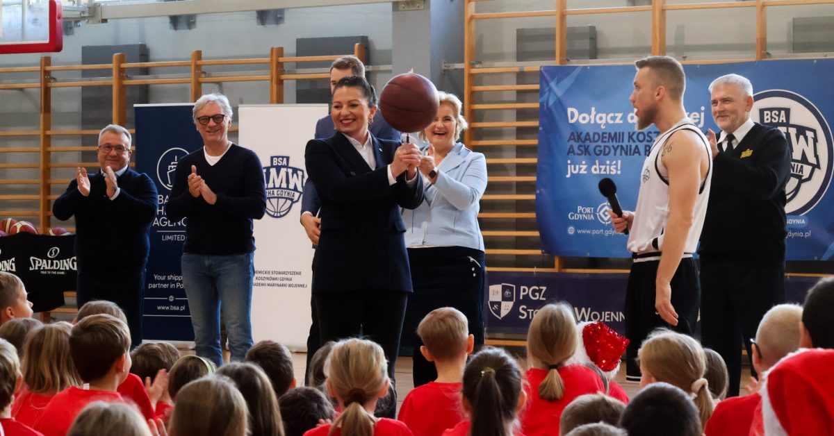 Pani w czarnym stroju trzyma piłkę do koszykówki na długopisie przed grupą dzieci w czerwonych koszulkach, obok stoją inni dorośli.