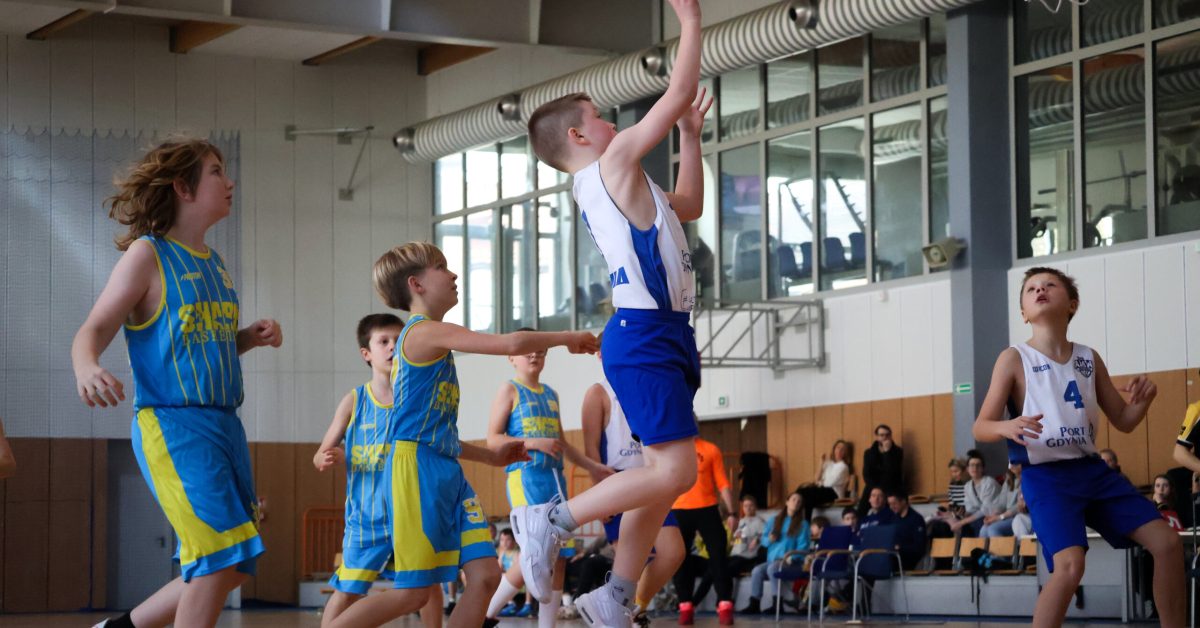 Chłopiec w białej koszulce wyskakuje do rzutu, podczas gdy przeciwnik w żółto-niebieskim stroju próbuje go dogonić.