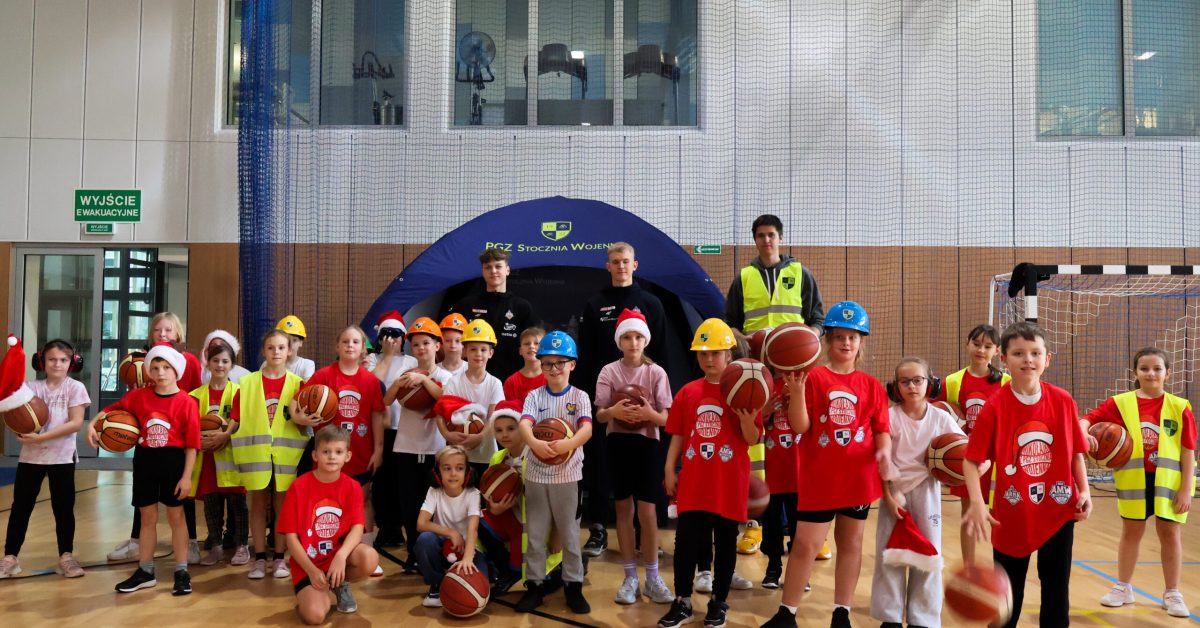 Grupa dzieci w kaskach budowlanych i kamizelkach odblaskowych pozuje do zdjęcia w hali sportowej.