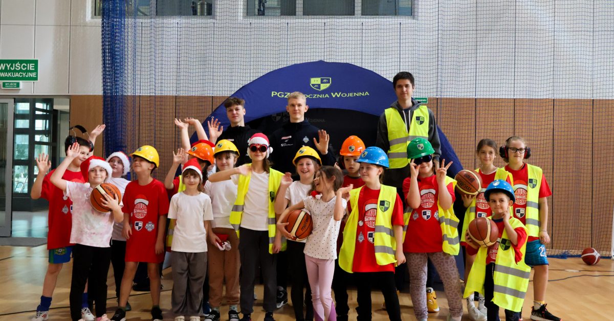 Grupa dzieci w kaskach budowlanych i kamizelkach odblaskowych pozuje do zdjęcia w hali sportowej.