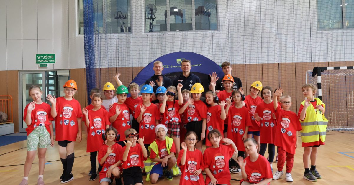 Grupa dzieci w kaskach budowlanych i kamizelkach odblaskowych pozuje do zdjęcia w hali sportowej.