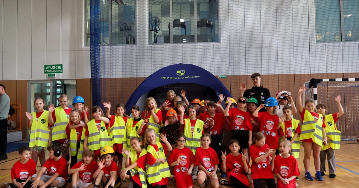 Grupa dzieci w kaskach budowlanych i kamizelkach odblaskowych pozuje do zdjęcia w hali sportowej.