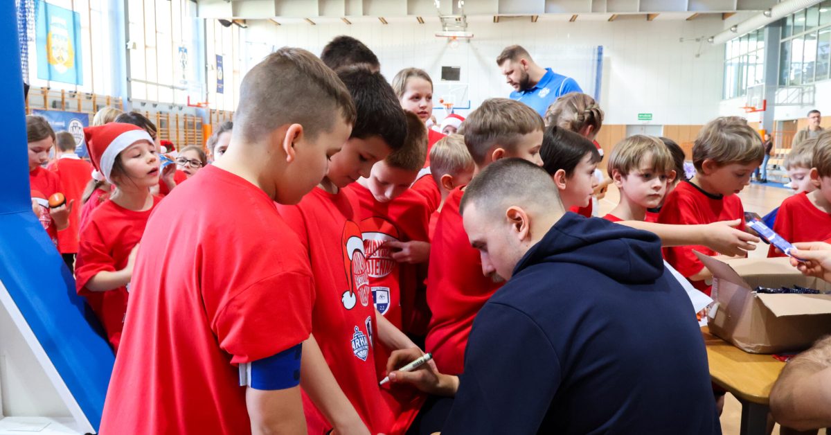 Dzieci w czerwonych koszulkach zbierają się wokół zawodnika ekstraklasy, który wręcza im autografy.