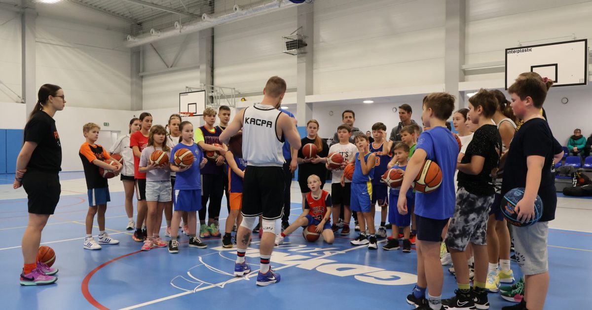 Kacpa w białej koszulce mówi do grupy dzieci stojących na niebieskim boisku koszykarskim.