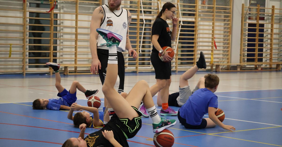 Zawodniczka w czarnej koszulce i czarnych spodenkach wykonuje ćwiczenie na podłodze, obok inni uczestnicy trenują, a nad nią Kacpa zdziwiony.