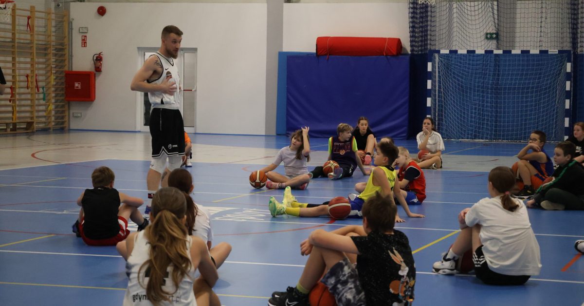 Dzieci siedzą na boisku, słuchając Kacpe podczas sesji treningowej.