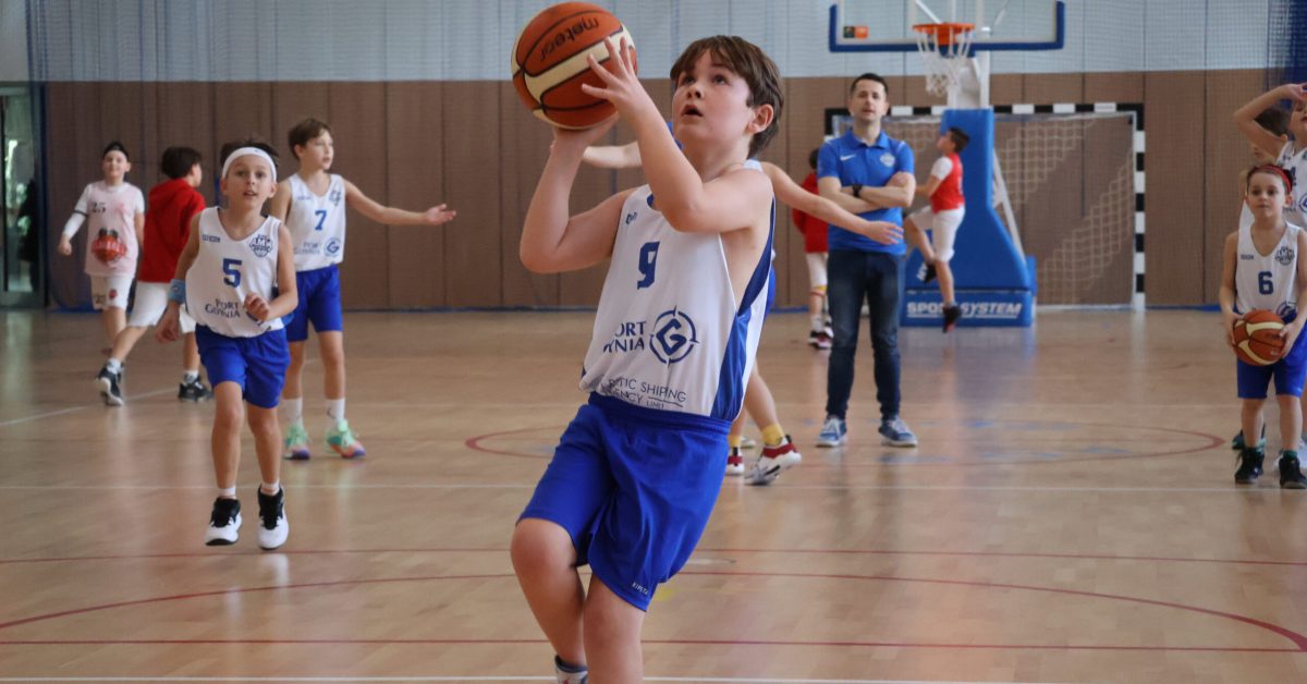 Chłopiec w białej koszulce skacze do rzutu, w tle pozostali zawodnicy na hali sportowej.