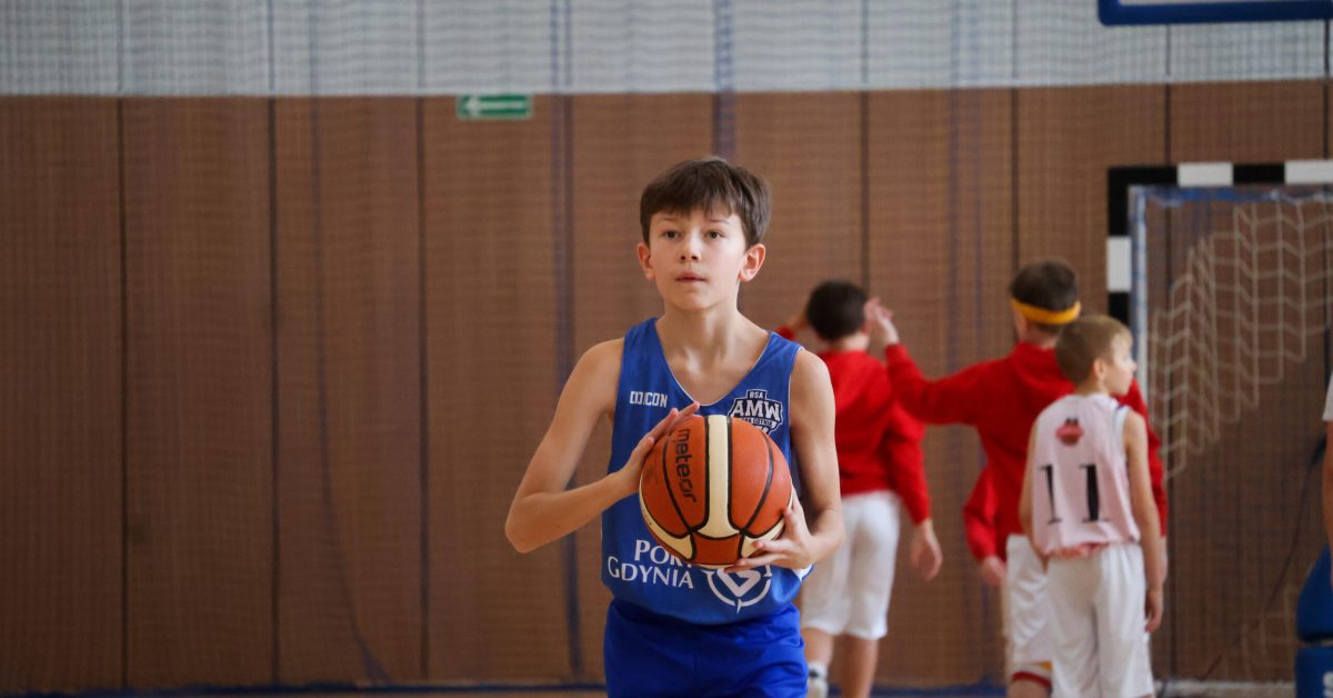 Chłopiec w niebieskiej koszulce trzyma piłkę do koszykówki i koncentruje się na rzucie, w tle inni zawodnicy.