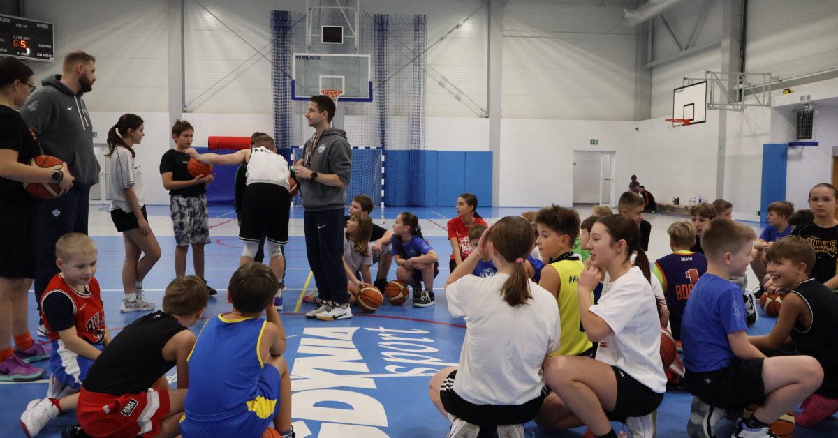 Grupa dzieci i młodzieży słucha trenera na środku boiska.