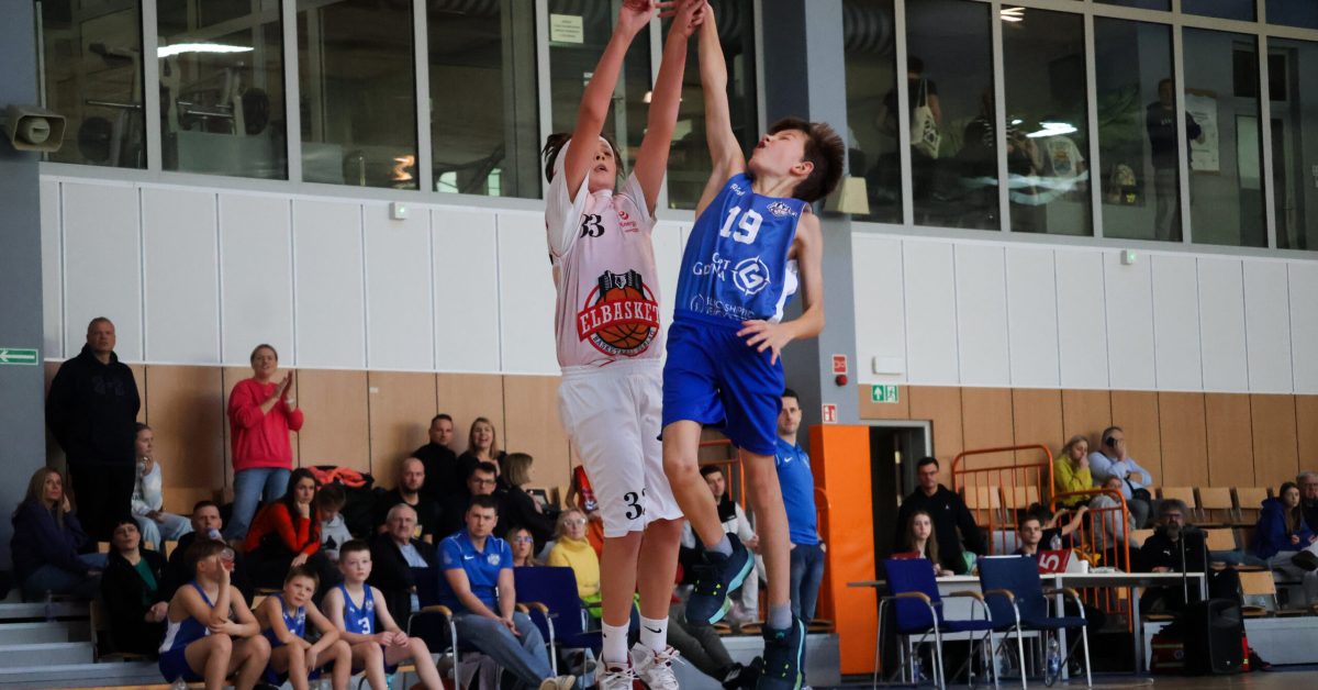 Zawodnik w białej koszulce próbuje zablokować rzut przeciwnika w niebieskim stroju, wyskakując w górę.