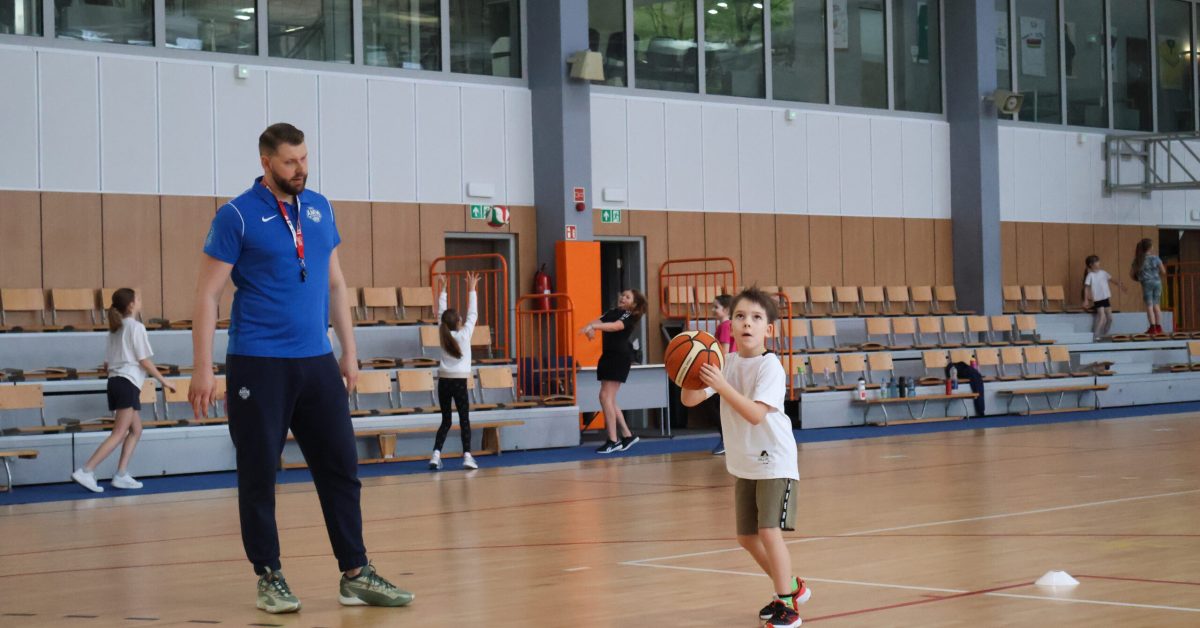 Trener w niebieskiej koszulce obserwuje młodego zawodnika w białej koszulce i zielonych spodenkach, który przygotowuje się do rzutu piłką.