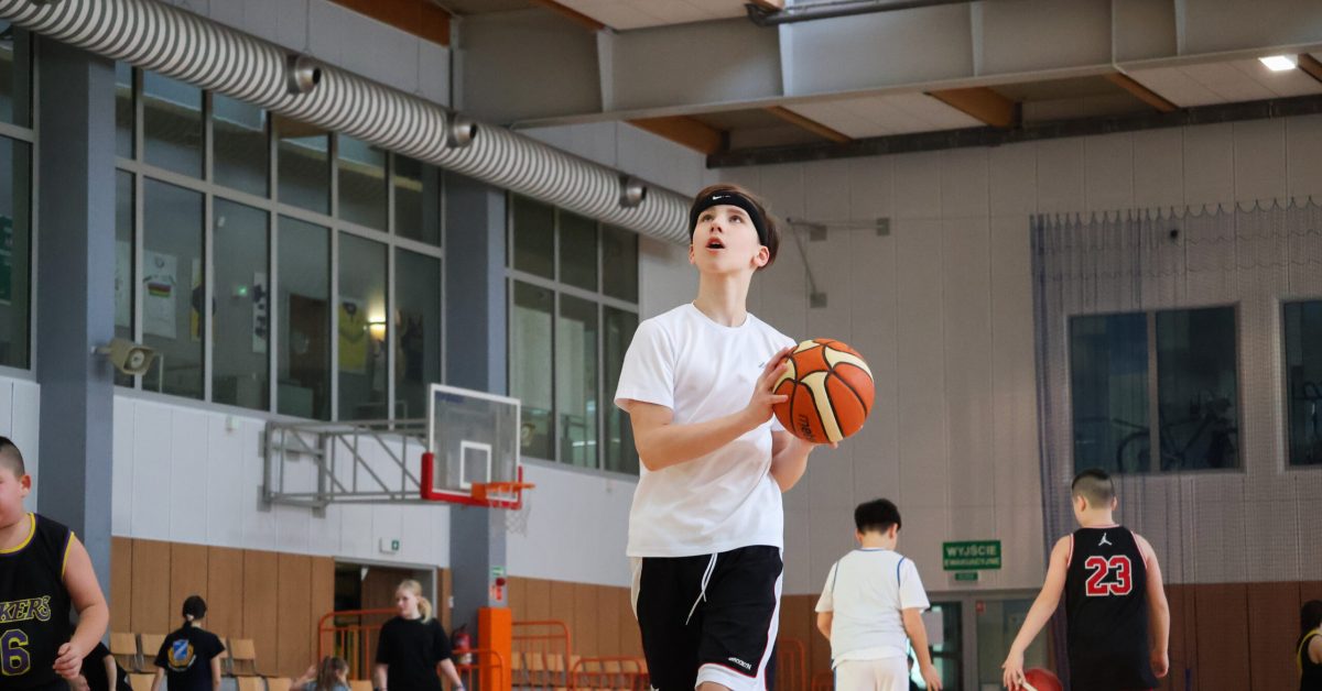 Chłopiec w białej koszulce i czarnych spodenkach wykonuje rzut do kosza na hali sportowej. W tle inni uczestnicy treningu.
