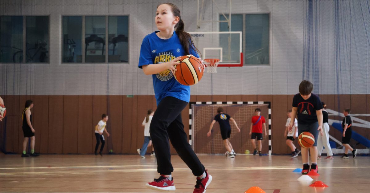 Dziewczynka w niebieskiej koszulce i czarnych spodniach biegnie po hali sportowej, trzymając piłkę do koszykówki.