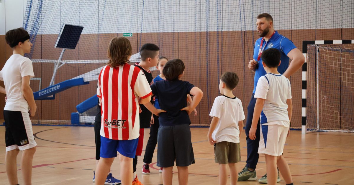 Grupa dzieci w koszulkach sportowych, w tym jedno w czerwono-białych pasach, słucha wskazówek trenera z gwizdkiem.