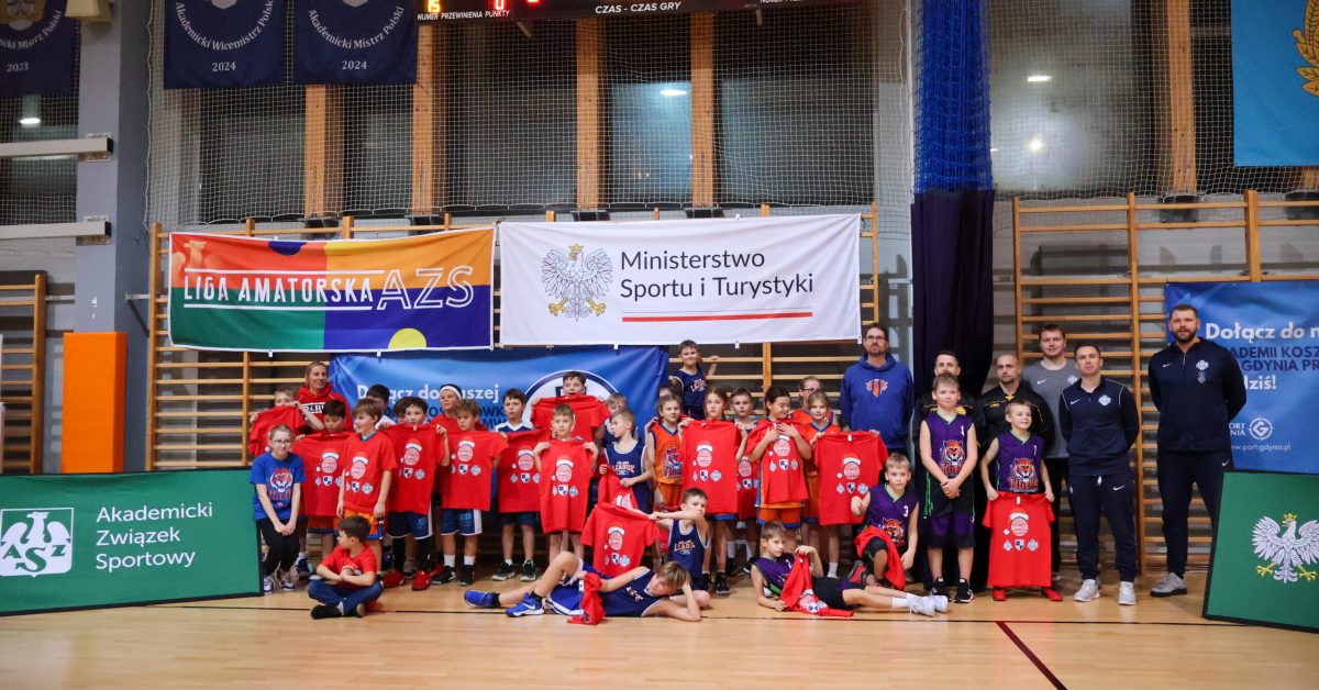 Zdjęcie grupowe uczestników turnieju koszykówki, zawodnicy i trenerzy ustawieni przed banerem sponsora.