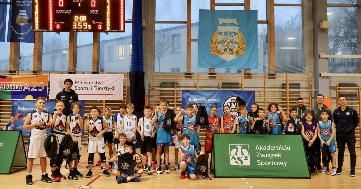 Zdjęcie grupowe uczestników turnieju koszykówki, zawodnicy i trenerzy ustawieni przed banerem sponsora.