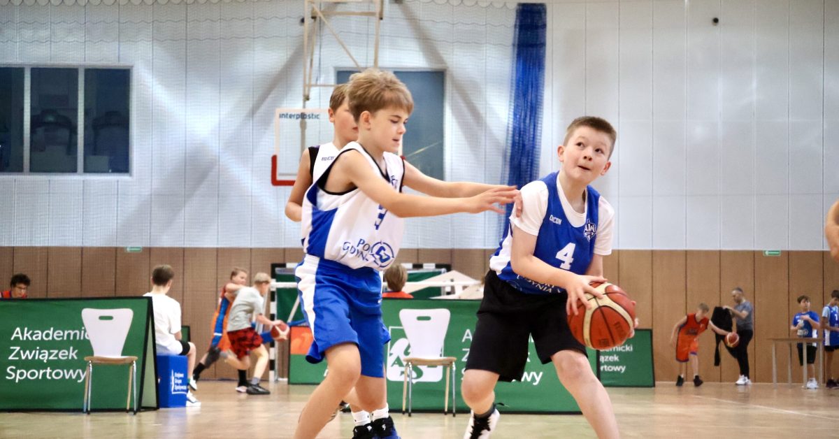 Chłopiec w niebieskiej koszulce z numerem oddaje rzut do kosza, a obrońca w białej koszulce próbuje go zablokować.