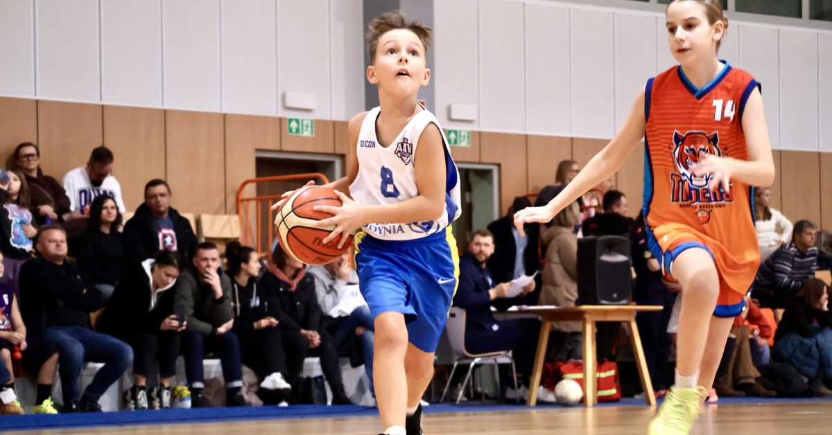 Młody zawodnik w niebiesko-białej koszulce prowadzi piłkę po boisku, przygotowując się do rzutu, w tle zapełnione trybuny.