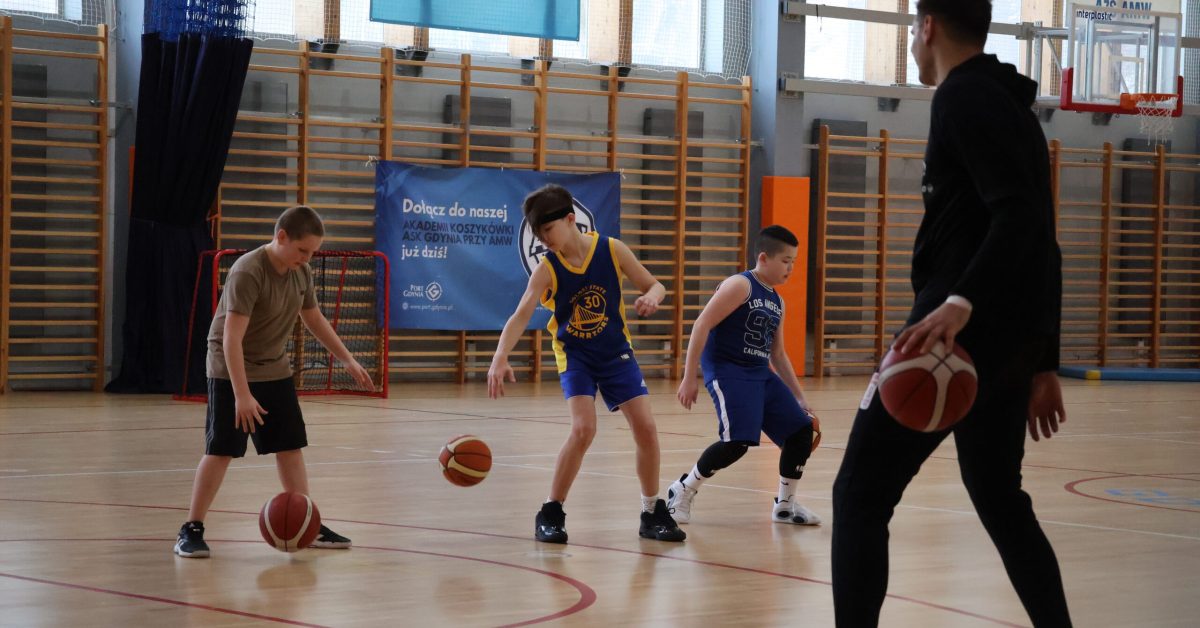 Trening koszykówki – kilku młodych zawodników drybluje piłkami, a zawodnik ekstraklasy Stefan Djordević obserwuje ich ruchy.