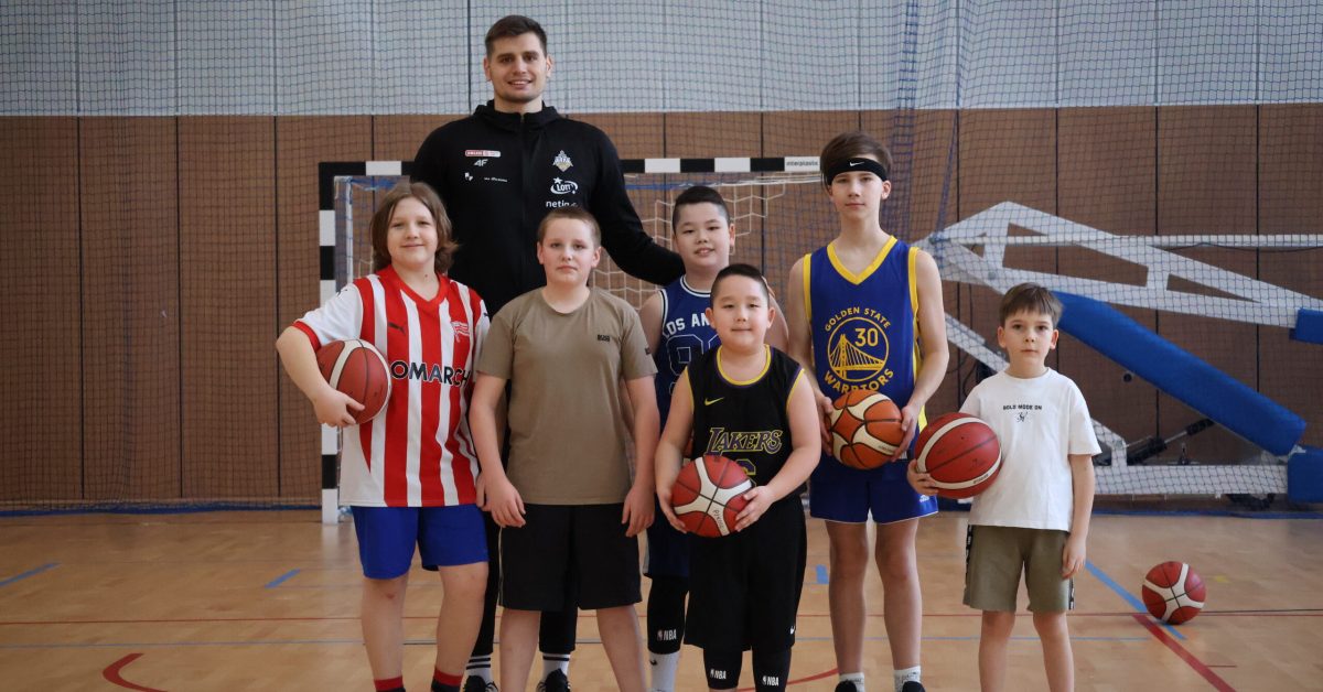 Grupa uśmiechniętych dzieci pozuje do zdjęcia na hali sportowej, za nimi stoi zawodnik ekstraklasy Stefan Djordevic.