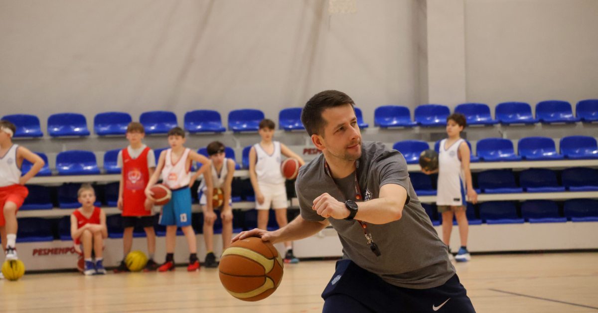 Trener w ciemnym stroju demonstruje ruch z piłką do koszykówki, stojąc przed grupą młodych zawodników. W tle widać ławkę i niebieskie trybuny oraz zawodników.