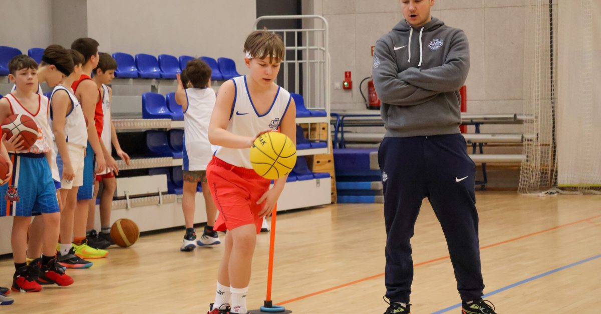 Trener w szarej bluzie i granatowych spodniach stoi z założonymi rękami, patrząc na chłopca w czerwonych spodenkach, który kozłuje żółtą piłkę między pachołkami.